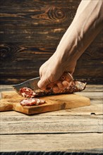 Hand with knife cutting smoked dried pork sausage on slices on wooden cutting board