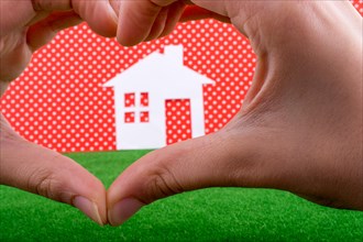 Paper house in the middle of a hand making a heart shape on a white-dotted red background with grass