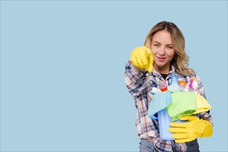 Young beautiful cleaner woman holding bucket with products pointing camera against blue backdrop. Resolution and high quality beautiful photo