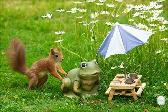 Squirrel leaning against frog next to table with potty and parasol in green grass and white flowers