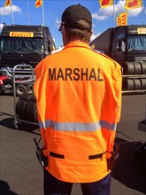 Track marshal of international car race is dressed in orange jacket with inscription Marshal on back carrying left radio
