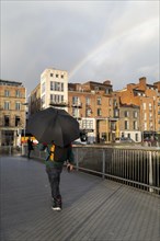 A man crosses a bridge in Dublin underneath an umbrella as a rainbow appears in the sky. Dublin