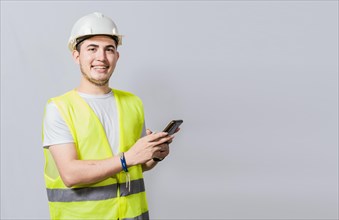 Smiling engineer using cell phone looking at camera. Friendly male builder engineer using phone and looking at camera. Young engineer in vest and helmet using cell phone smiling at camera