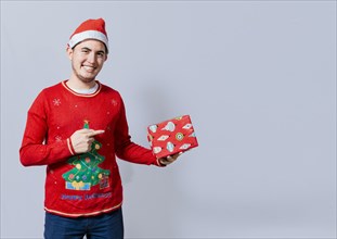 Smiling guy in christmas hat holding and pointing a gift box. Portrait of teenager in christmas hat holding and pointing a gift box. Guy in christmas hat and sweater holding a gift box isolated