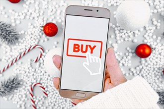 Concept for Christmas seasonal gift online shopping with hand holding cell phone Buy button on display in front of background with white seasonal decoration