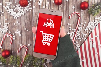 Concept for Christmas seasonal online gift shopping with hand holding cell phone with white shopping cart sign on red background in front of wooden desk with seasonal decorations