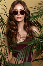 Beautiful sexy woman in a bikini and sunglasses posing in the middle of green plants. Summer look. Beauty face. Photo taken in the studio