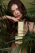 Beautiful sexy woman in a bikini posing in the middle of green plants with a box of cosmetics in hand. Summer look. Beauty face. Photo taken in the studio