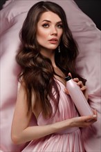 Beautiful brunette woman with curls and classic make-up in a flying light pink dress with a bottle of shampoo in hands. Beauty face. Photo taken in studio