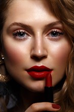 Beautiful girl with red lips and classic makeup and curls with lipstick in hand. Beauty face. Photo taken in the studio