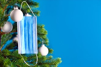 Medical face mask hanging from Christmas tree on blue background with empty copy space. Concept for Christmas celebration during times of Corona virus