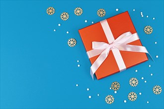 Christmas gift box with ribbon surrounded by seasonal wooden snowflake ornaments and white snowballs on side of blue background with empty copy space