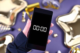 New Year Silvester celebration concept with hand holding smart phone with timer countdown to midnight in front of party items in background