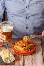 Man in blue shirt eating a clay pot with meatballs and potatoes and a beer in his hand