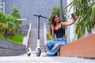 Portrait of a young woman sitting in the city with an electric scooter and taking a selfie