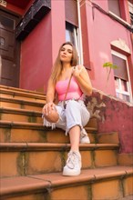 Young blonde caucasian tourist sitting on the stairs of a street with houses with colorful facades