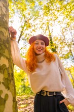 Red-haired woman leaning against a tree in a forest park smiling at sunset