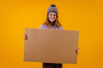 Woman in a wool cap holding a cardboard sign on a yellow background