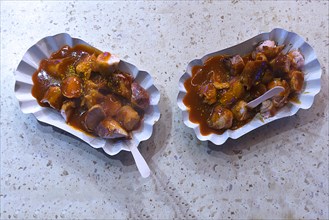 Two curry sausage portions in cardboard bowls with wooden fork at a sausage stand