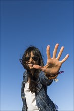 Low angle view of a similing woman while showing the palm of her hand. Blue sky background. Stop gesture