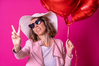 Blond caucasian woman in a white hat and sunglasses making the victory sign with her hands in a nightclub with some heart balloons