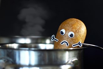 Potato with sad face and goggle eyes being put into a steaming cooking pot on dark black background
