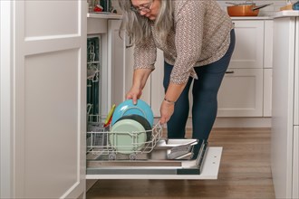 Older white-haired woman putting dishes in the dishwasher in her home kitchen