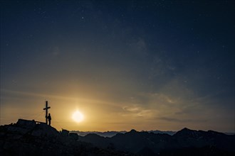 Mountaineer at summit cross of Namloser Wetterspitze at full moon and Lechtaler Alps