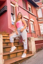 Young blonde caucasian woman sitting on the stairs of a street with houses with colorful pink facades