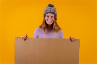 Portrait of a woman in a wool cap holding a cardboard sign on a yellow background