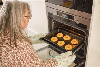 Older white-haired woman putting a tray of cookies in the oven