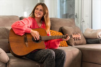 Woman playing the guitar at home smiling sitting on the sofa in her living room next to the sale