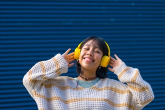 Asian girl listening to music in yellow headphones with her eyes closed on a blue college background