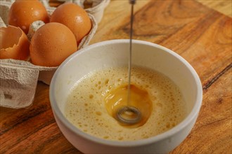Beating an egg in a bowl with a small mixer with an egg cup at the bottom on a wooden table