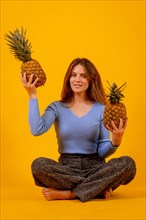 Portrait of smiling vegetarian woman with a cut pineapple sitting on a yellow background