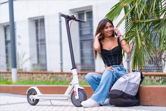 Young woman sitting in the city waiting for friends with an electric scooter talking on the phone