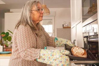 Older white-haired woman putting bread in the oven in her kitchen at home