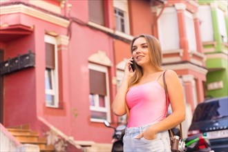 Young blonde caucasian woman in a street with houses with pink colorful facades with the phone