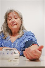 Older white-haired woman with glasses taking her blood pressure at home with white background