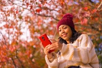 Asian girl in autumn with a mobile writing a message in a forest of red leaves