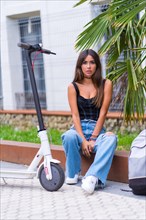 Portrait of a young caucasian woman sitting in the city waiting for friends with an electric scooter