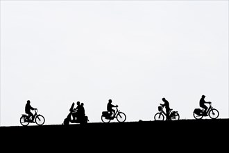 Motor scooter riding past cyclists and man on bicycle checking his smartphone in summer silhouetted against white background