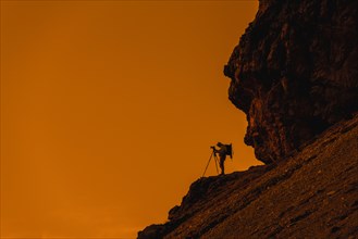 Landscape photographer in cliff face taking nature pictures of mountains with tripod at sunset in the Dolomites