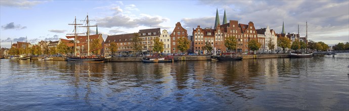 A panoramic photo of historic sailing ships on the banks of the river Trave and behind them the brick buildings of the old town