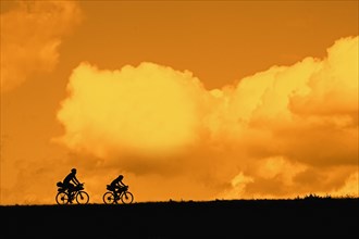 Couple of young cyclists riding their touring bicycles silhouetted against orange sunset sky in summer