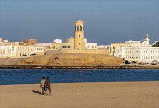 Two men walk on Sur Beach with Al-Ayjah Lighthouse in the background