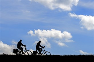 Two elderly cyclists silhouetted riding their bicycles against cloudy sky in summer