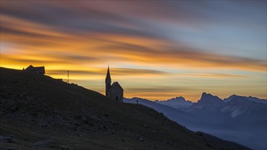 Little chapel silhouetted against sunset and view over Dolomite mountain range in summer