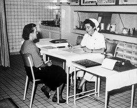 Old black and white archival photograph showing female factory worker visiting nurse at dispensary in the 1950s
