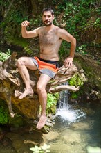 Man in swimsuit sitting on a tree trunk on a river bank